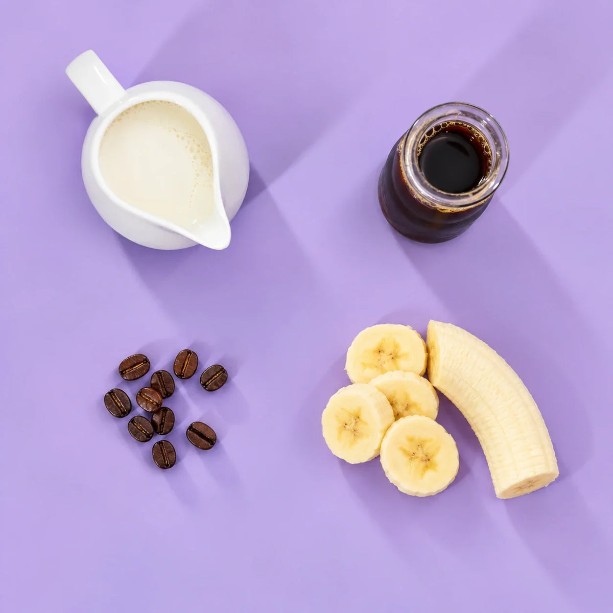 Iced coffee banana smoothie ingredients: chilled coffee, banana, coffee beans and milk on a lavender background
